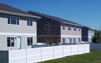 A row of beige townhouses behind a white fence on a sunny day.